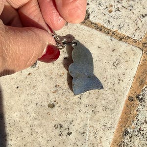 May include: A silver-toned pendant in the shape of a whale tail. The pendant has a blue and gray color scheme. The pendant is attached to a silver chain. The background is a light-colored, textured surface.