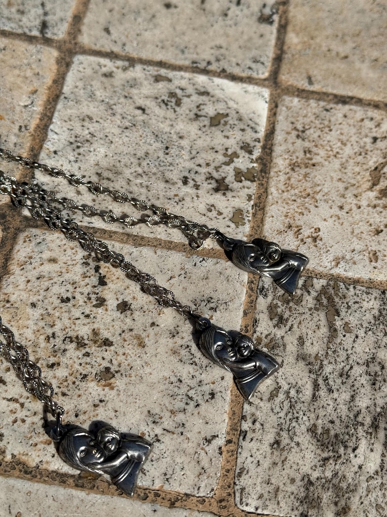 May include: Three silver-toned necklaces with pendant charms. Each charm depicts a mother and child in a loving embrace. The chains have a delicate, twisted design. The necklaces are displayed on a textured, neutral-toned surface.