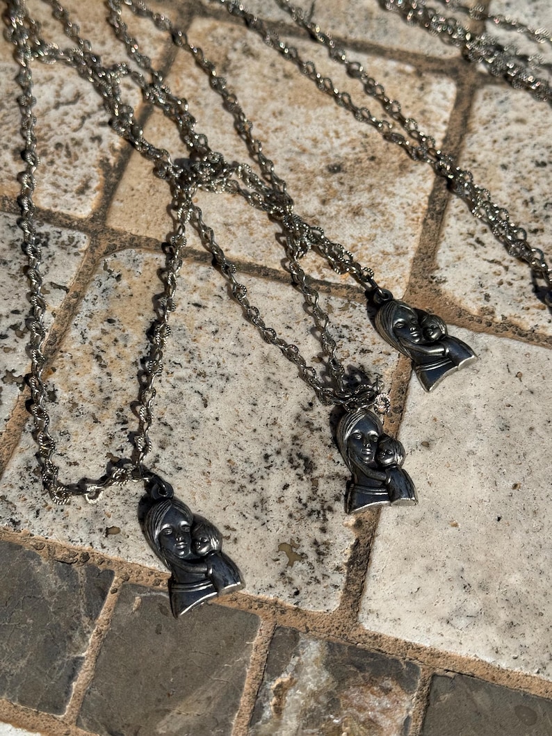 May include: Silver-toned necklaces featuring a pendant depicting a mother and child. The pendants are attached to delicate, twisted rope chains. The necklaces are displayed on a textured, stone-like surface.