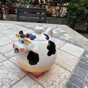 Op de afbeelding: Een witte theepot van keramiek in de vorm van een koe met zwarte vlekken. De theepot heeft een deksel en een tuit. Het gezicht van de koe is geschilderd met bruine ogen en een roze neus. De theepot staat op een betegelde ondergrond.