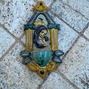 May include: A ceramic wall plaque showing a mother and child. The artwork uses blue, yellow, and green. The mother is holding the child. The piece has an arched top and decorative elements. The measurements are in centimetres.