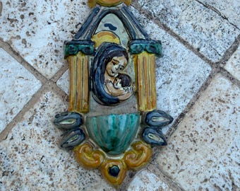 May include: A ceramic wall hanging depicting a mother and child. The artwork features a color palette of blue, yellow, and green. The mother is cradling the child. The piece has an arched top and decorative elements.