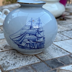 Puede incluir: Un jarrón redondo de cerámica azul claro con borde dorado. El jarrón presenta una ilustración azul detallada de un velero. El jarrón está sobre una superficie de piedra.