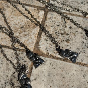 May include: Silver-toned necklaces with a pendant depicting a mother and child. The pendants are detailed with facial features and the chains have a delicate, linked design. The necklaces are displayed on a textured, stone-like surface.