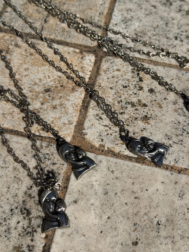 May include: Silver-toned necklaces with a pendant depicting a mother and child. The pendants are detailed with facial features and the chains have a delicate, linked design. The necklaces are displayed on a textured, stone-like surface.