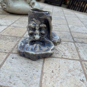 Op de afbeelding: Een keramiek beeldje van een baardig mannengezicht met een blauwe en witte glazuur. Het beeldje is gevormd als een kleine pot met een brede opening aan de bovenkant.