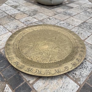 May include: A circular brass tray with detailed etched patterns, including a central star design and floral motifs. The tray sits on a stone surface. The decorative border and overall design suggest an antique or vintage aesthetic.