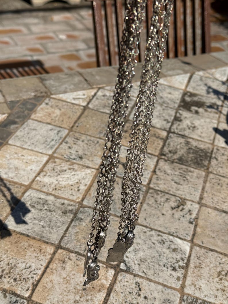 May include: Silver-toned, layered necklaces with small, decorative charms at the bottom. The chains are delicate and hang vertically against a neutral, textured background. The charms appear to be floral or leaf-shaped.