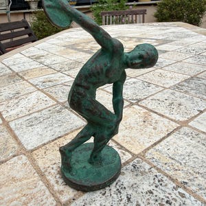 May include: A bronze statue of a discus thrower in mid-motion. The sculpture has a weathered, green patina, with the figure's arm extended, holding the discus. The statue is mounted on a circular base, set on a stone patio.