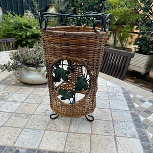 May include: A brown wicker umbrella stand with a black metal frame and decorative leaf cutouts. The stand is on a tiled surface.