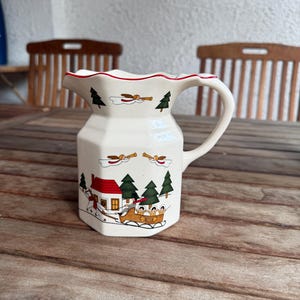 Può includere: Brocca in ceramica color crema con bordo smerlato e manico. La brocca è decorata con una scena natalizia con una casa con tetto rosso, alberi sempreverdi e una slitta con passeggeri. Sono raffigurati anche angeli con trombe.