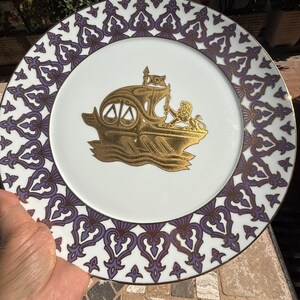 Peut inclure: Assiette en céramique blanche avec un emblème doré d'un navire et d'un lion. L'assiette a une bordure à motifs violets et blancs. L'assiette est tenue par une main.