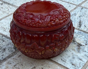 May include: A round, deep red lidded container featuring intricate, raised relief carvings of various fruits, including grapes and berries, along with decorative leaves and bow motifs covering the entire exterior surface and the lid.