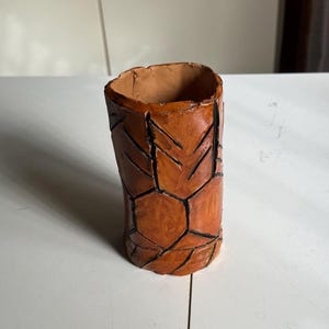 Può includere: Un vaso in ceramica marrone con un motivo geometrico inciso sulla superficie. Il vaso ha una forma cilindrica e un bordo leggermente svasato.