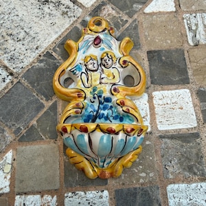 May include: A decorative ceramic wall fountain with a yellow, blue, and white color scheme. The fountain features a basin with a floral design and a top section with two cherubic figures. The piece is set against a stone tile background.