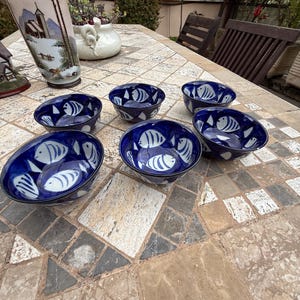 May include: Six blue ceramic bowls with a white fish design are arranged on a stone table. The bowls have a deep blue exterior and a white interior with a fish pattern. Other decorative items are in the background.