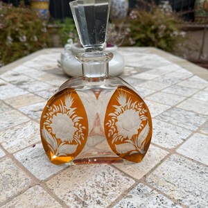 May include: A clear glass perfume bottle with an amber-colored design. The bottle features a clear, faceted stopper and etched floral designs on the front. The bottle is sitting on a tiled surface.