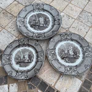 Puede incluir: Tres platos decorativos antiguos con un diseño de paisaje en blanco y negro. Cada plato presenta una escena central con figuras, urnas y elementos arquitectónicos, rodeada por un borde detallado. Los platos están dispuestos sobre una superficie estampada.