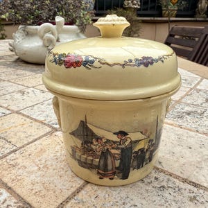 Puede incluir: Un tarro de cerámica vintage con tapa, que presenta una escena pintada a mano de personas en un mercado. El tarro tiene una base de color crema con detalles florales y una escena detallada de personas con ropa tradicional.