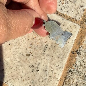 May include: A small, silver-colored pendant with a stylized Egyptian pharaoh design. The pendant is held between fingers, showcasing its shape and the attached loop for a chain. The surface of the pendant has a slightly worn appearance.