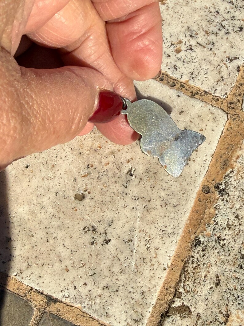 May include: A small, silver-colored pendant with a stylized Egyptian pharaoh design. The pendant is held between fingers, showcasing its shape and the attached loop for a chain. The surface of the pendant has a slightly worn appearance.