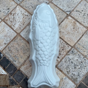 May include: A white ceramic fish-shaped mold with detailed scales, fins, and a visible eye. The mold is designed for baking or decorative purposes. The fish is displayed on a tiled surface.