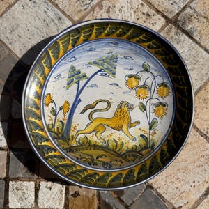 May include: A round ceramic plate with a blue, green, and yellow border. The center of the plate features a yellow lion with a blue background and a green and yellow floral design.