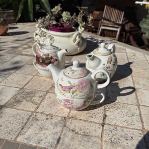 Puede incluir: Tres juegos de té individuales de cerámica, cada uno compuesto por una tetera apilada sobre una taza a juego. Los juegos presentan patrones florales, de pájaros y mariposas en tonos rosa, verde y dorado. Un juego muestra una ilustración de un gato.