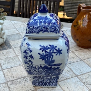May include: A blue and white porcelain jar with a lid. The jar features a floral design and a geometric pattern around the base. The jar is sitting on a tiled surface.