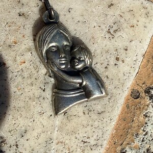 May include: Silver-toned pendant necklace featuring a mother and child embracing. The pendant depicts a woman holding a child close, with detailed facial features and flowing hair. The necklace chain is also silver-toned.
