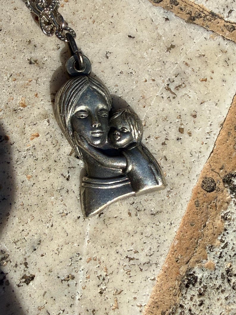 May include: Silver-toned pendant necklace featuring a mother and child embracing. The pendant depicts a woman holding a child close, with detailed facial features and flowing hair. The necklace chain is also silver-toned.