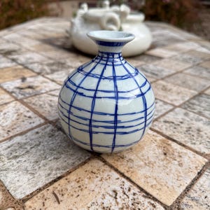 May include: A small, round, white ceramic vase with a blue grid pattern. The vase has a narrow neck and a slightly flared rim. It is sitting on a stone tile surface.