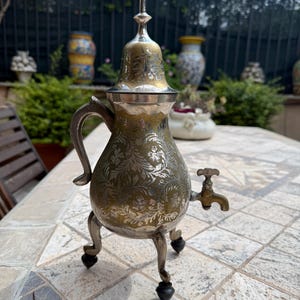 Op de afbeelding: Een metalen vat met een bolvormig lichaam, voorzien van een gedetailleerde zilverkleurige bloemengravure op een dof goudkleurig oppervlak. Het heeft een gebogen handvat, drie slanke poten met donkere voetjes, een puntig deksel en een kleine kraan.