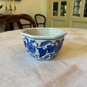 Può includere: Una piccola ciotola in ceramica bianca con un motivo floreale blu. La ciotola ottagonale presenta un disegno di fiori e foglie blu. Il bordo è semplice. Questa ciotola decorativa è probabilmente per l'esposizione o piccoli oggetti.