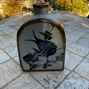 Puede incluir: Un jarrón de cerámica con un diseño de pez azul y blanco. El jarrón está decorado con acentos florales verdes y azules. El jarrón tiene un borde marrón y una base marrón.