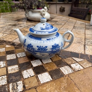 Puede incluir: Una tetera pequeña de color blanco con un diseño floral azul y blanco. La tetera tiene una tapa y un pico. Está sentada sobre una superficie de azulejos.
