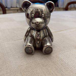 May include: A silver teddy bear figurine with a buttoned shirt and a shiny finish.