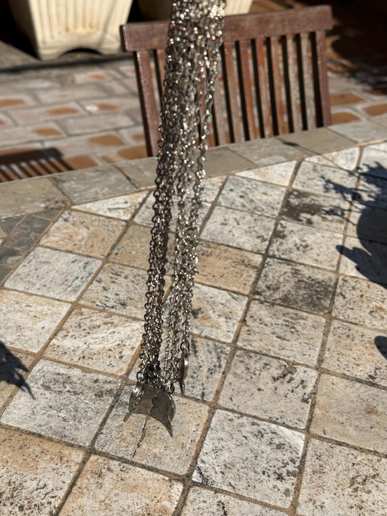 May include: Silver-toned necklace with multiple strands and leaf-shaped pendants. The necklace is hanging, showcasing its intricate chain design and delicate charms. The jewelry is displayed against a neutral background, highlighting its metallic sheen.