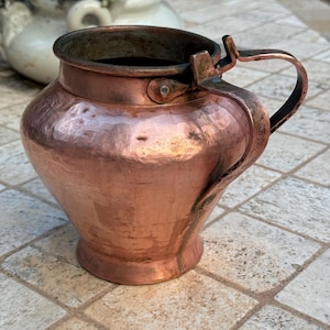 Puede incluir: Una jarra de cobre con un acabado martillado y un asa. La jarra tiene un cuerpo redondeado y un labio ligeramente acampanado.