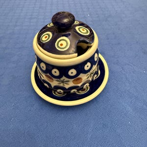 Può includere: Una zuccheriera in ceramica con coperchio e piattino abbinati, caratterizzata da un motivo blu scuro, crema e marrone con cerchi e forme a stella. Il coperchio presenta un piccolo pomello e una tacca per il cucchiaino, con finitura lucida.