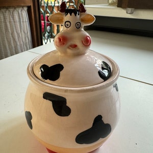 May include: A ceramic cookie jar shaped like a cow with black and white spots. The lid is shaped like a cow's head with pink cheeks and black eyes. The jar has a red base.