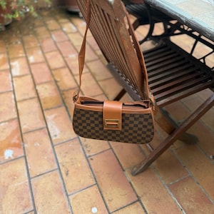 Peut inclure: Un sac à bandoulière marron et beige à carreaux, de forme rectangulaire et avec une fermeture à rabat. Le sac a une bandoulière en cuir marron et une boucle dorée. Le sac est suspendu à une chaise en bois.