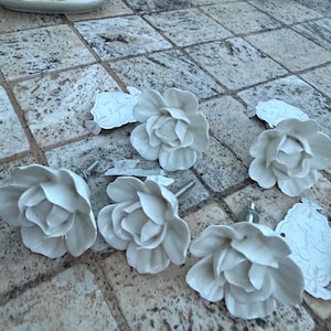 Peut inclure: Boutons de meubles blancs en céramique en forme de fleur avec des fixations argentées. Les boutons sont disposés sur une surface en dalles de pierre. Des étiquettes décoratives sont également présentes.