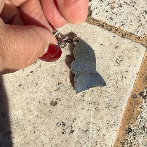 May include: A silver necklace with a pendant in the shape of a fish. The pendant is a muted blue-grey color. The necklace is held by a hand with red nail polish against a textured, light-colored surface.