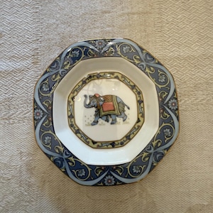 Peut inclure: Une assiette décorative octogonale avec une bordure florale bleue et dorée. Le centre présente un motif d'éléphant avec une couverture rouge et orange. L'assiette est blanche avec un bord doré.