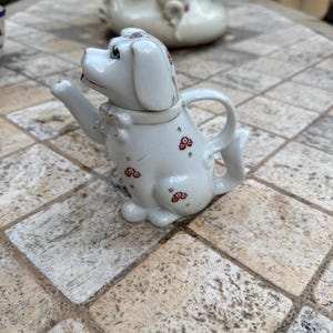 Könnte beinhalten: Weiße Porzellan-Teekanne in Form eines Hundes mit roten und goldenen floralen Akzenten. Die Teekanne hat einen Ausguss und einen Griff.