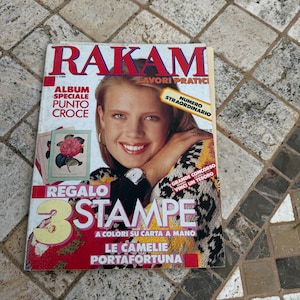 May include: A magazine cover with the title "Rakam" in red letters. The cover features a woman with blonde hair wearing a colorful sweater. The text "Regalo 3 Stampe" is in a large font and the text "A colori su carta a mano Le Camelie Portafortuna" is in a smaller font.