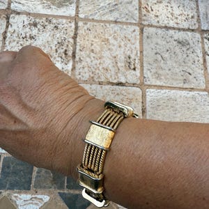 May include: Gold-toned bracelet featuring a woven design with rectangular and curved accents. The bracelet is composed of multiple strands of gold-colored metal, creating a textured appearance. The clasp is a gold-colored hook and eye closure.