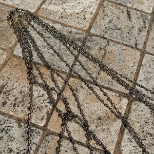 May include: Multiple strands of silver-toned necklaces with pendant charms. The chains are draped across a textured, neutral-toned surface. The pendants feature a detailed face design.