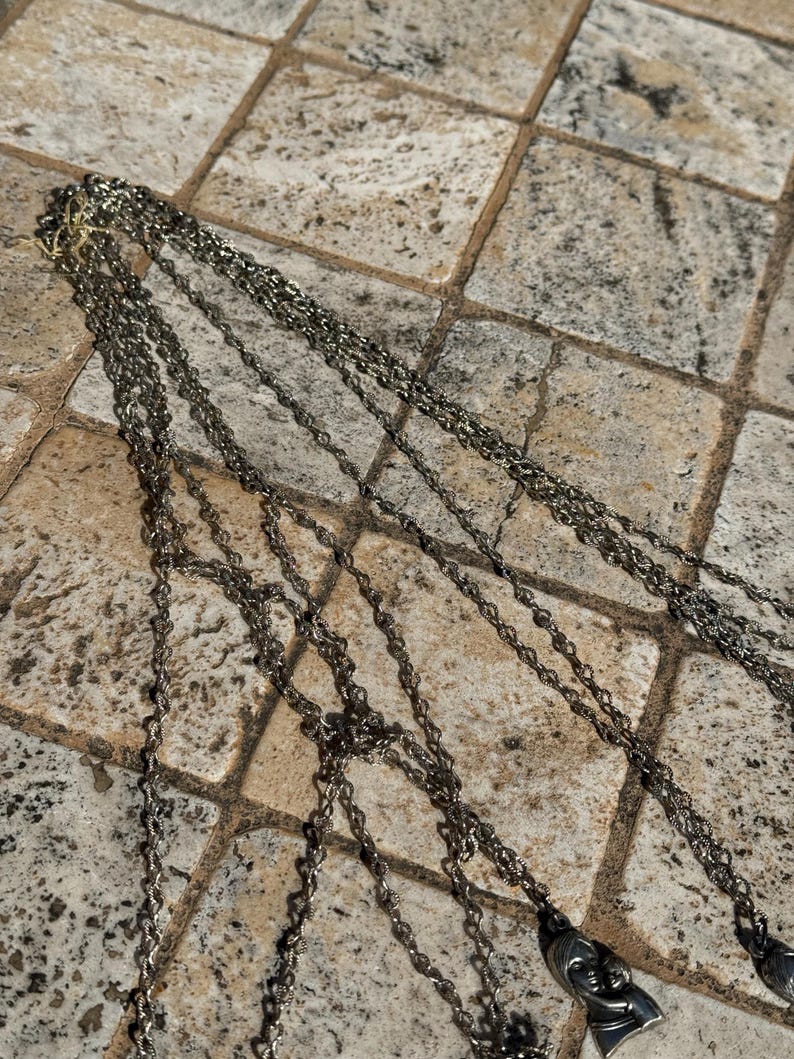 May include: Multiple strands of silver-toned necklaces with pendant charms. The chains are draped across a textured, neutral-toned surface. The pendants feature a detailed face design.